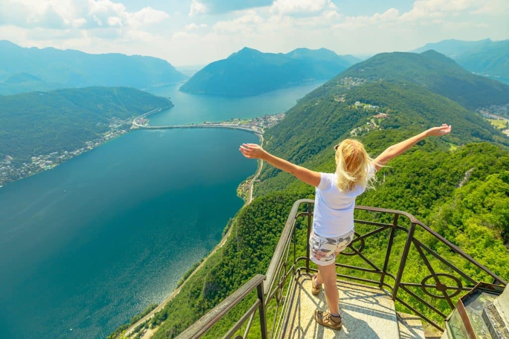 Swiss woman with open arms on top of San Salvatore mount by Lugano Lake in Switzerland. Aerial view lookout of Lugano with in Ticino canton.